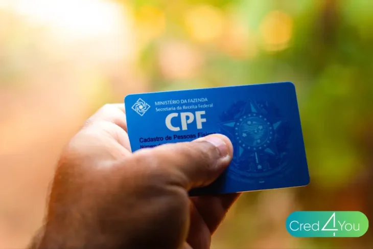 Pessoa segurando um cartão azul do CPF em primeiro plano, com fundo desfocado; imagem relacionada ao tema consultar CPF grátis.