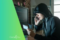 Pessoa com capuz usando computador e segurando cartão bancário ao telefone, representando fraudes financeiras e crimes cibernéticos.