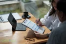 Beratung zur Pflegeversicherung, zwei Personen prüfen Unterlagen auf einem Klemmbrett am Tisch mit Laptop.